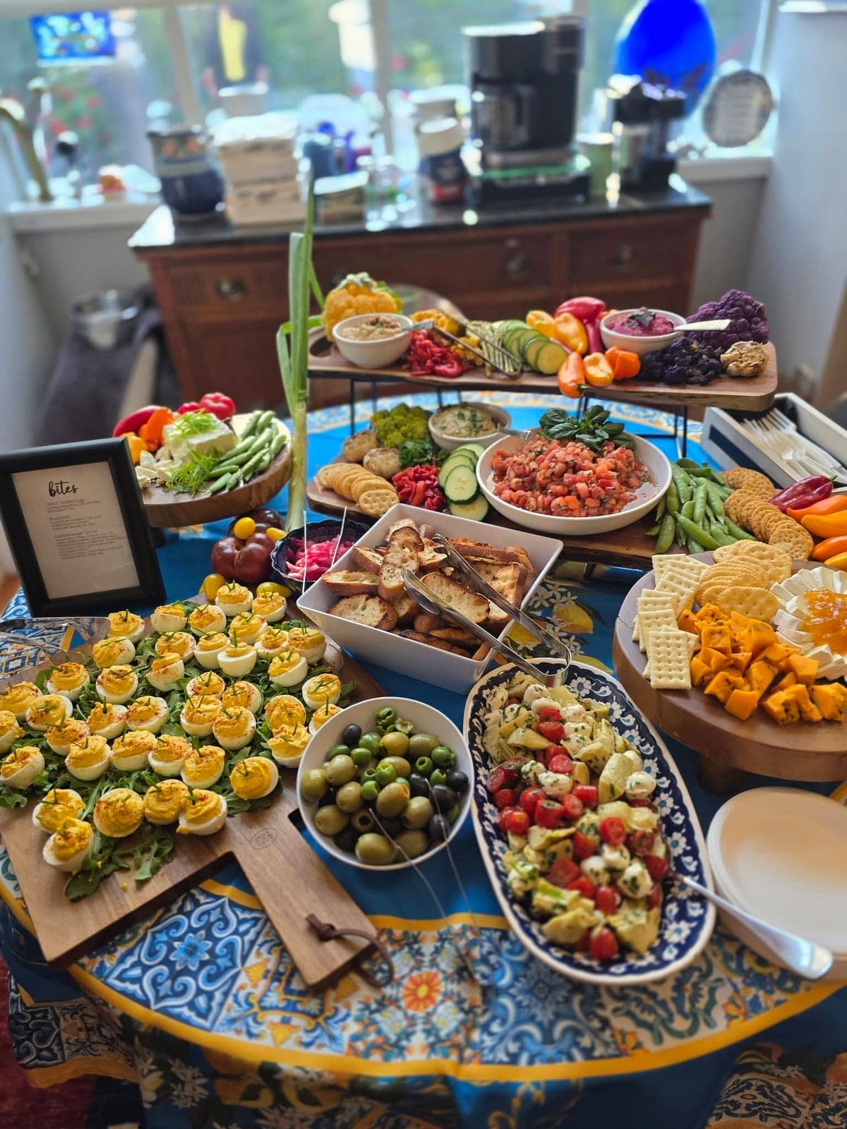A spread on a patterned tablecloth with deviled eggs, fruit, cheeses, olives and serving platters.