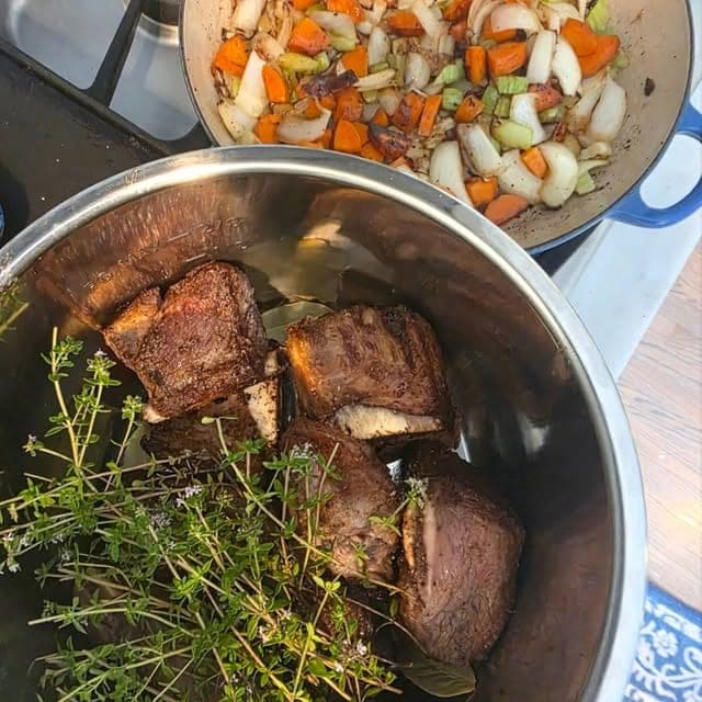 Seared shanks in a pot with thyme, beside a pan of diced vegetables.