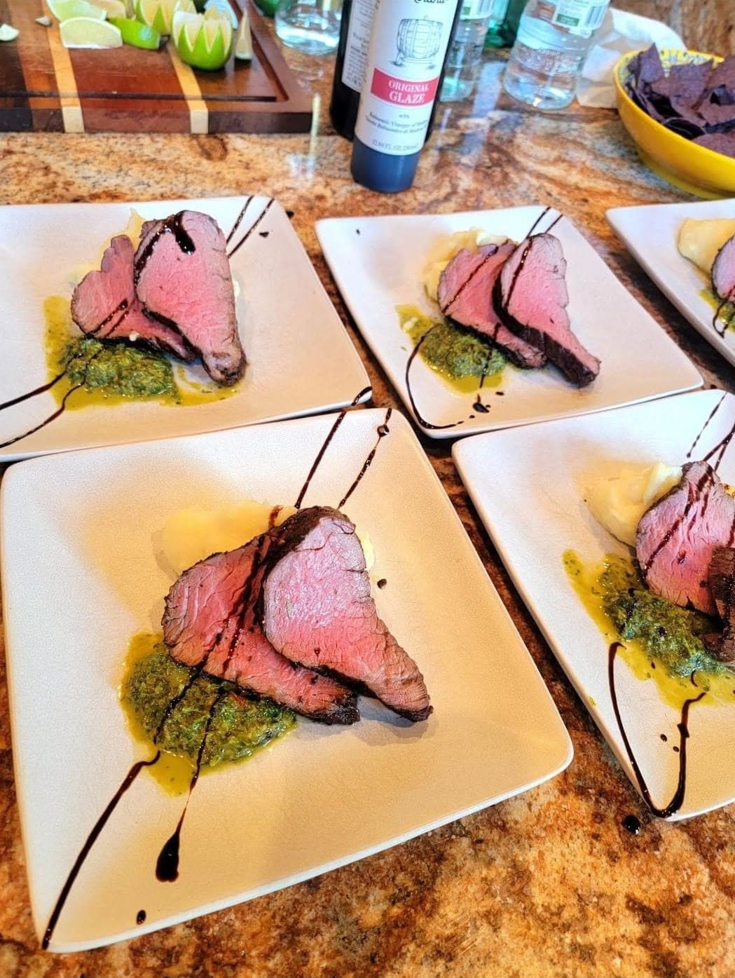 Several square white plates lined up, each with slices of rare seared beef tenderloin over a green herb sauce with a balsamic drizzle.